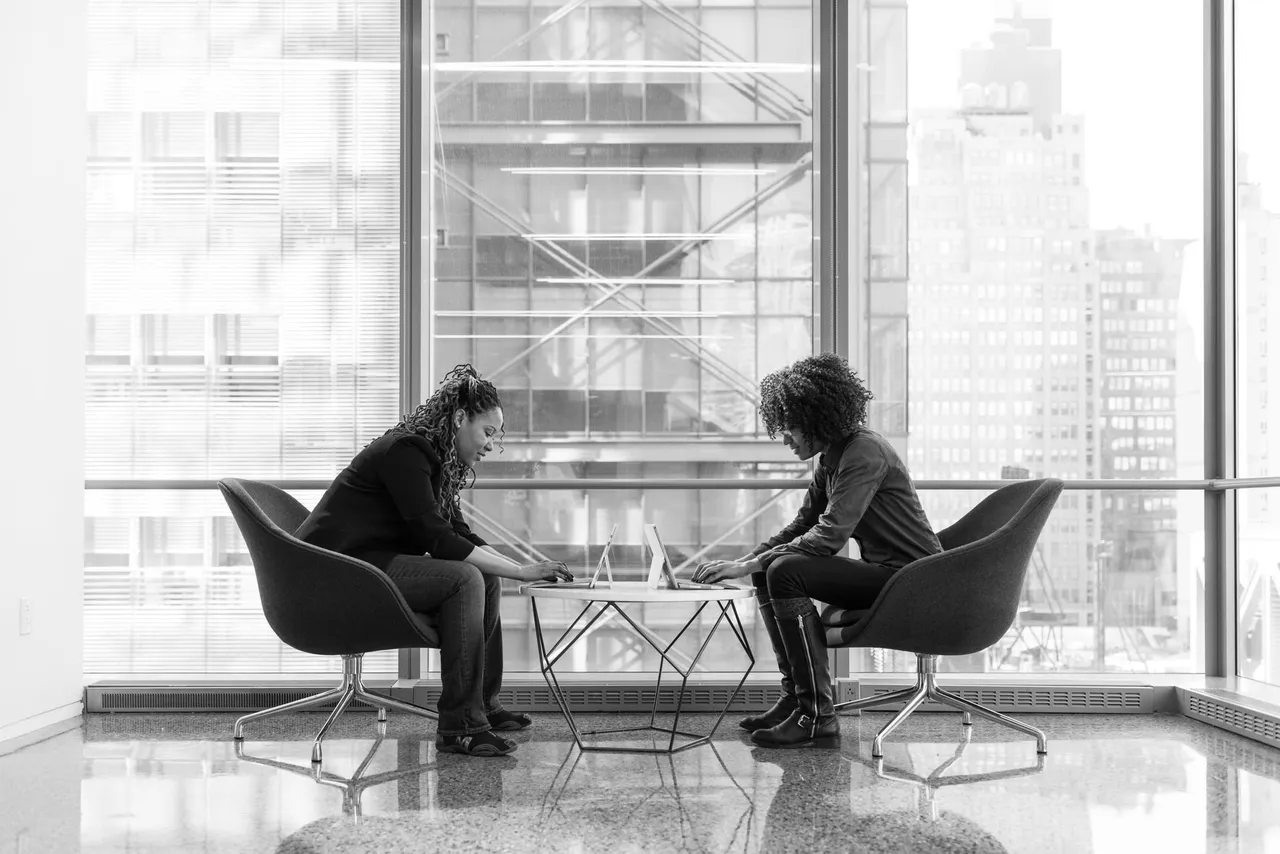 Two women talking across a table in a modern office with a city view.