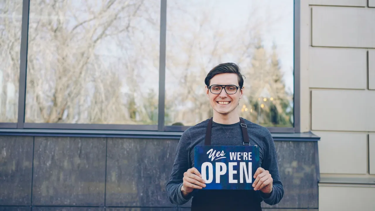 Smiling young business owner in glasses holding 'Yes, we're open' sign in front of store.