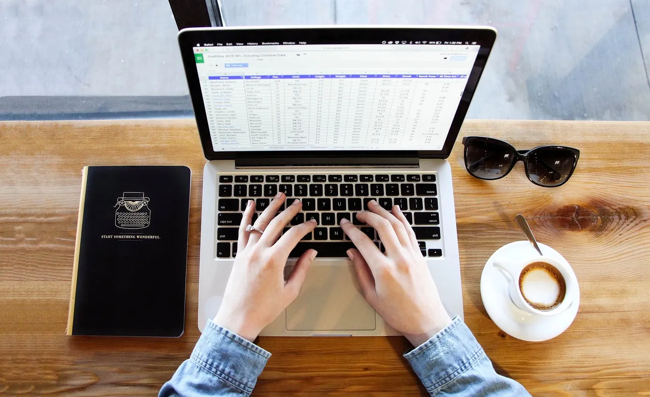 Hands typing on a laptop with a spreadsheet open, next to a coffee cup and a notebook.