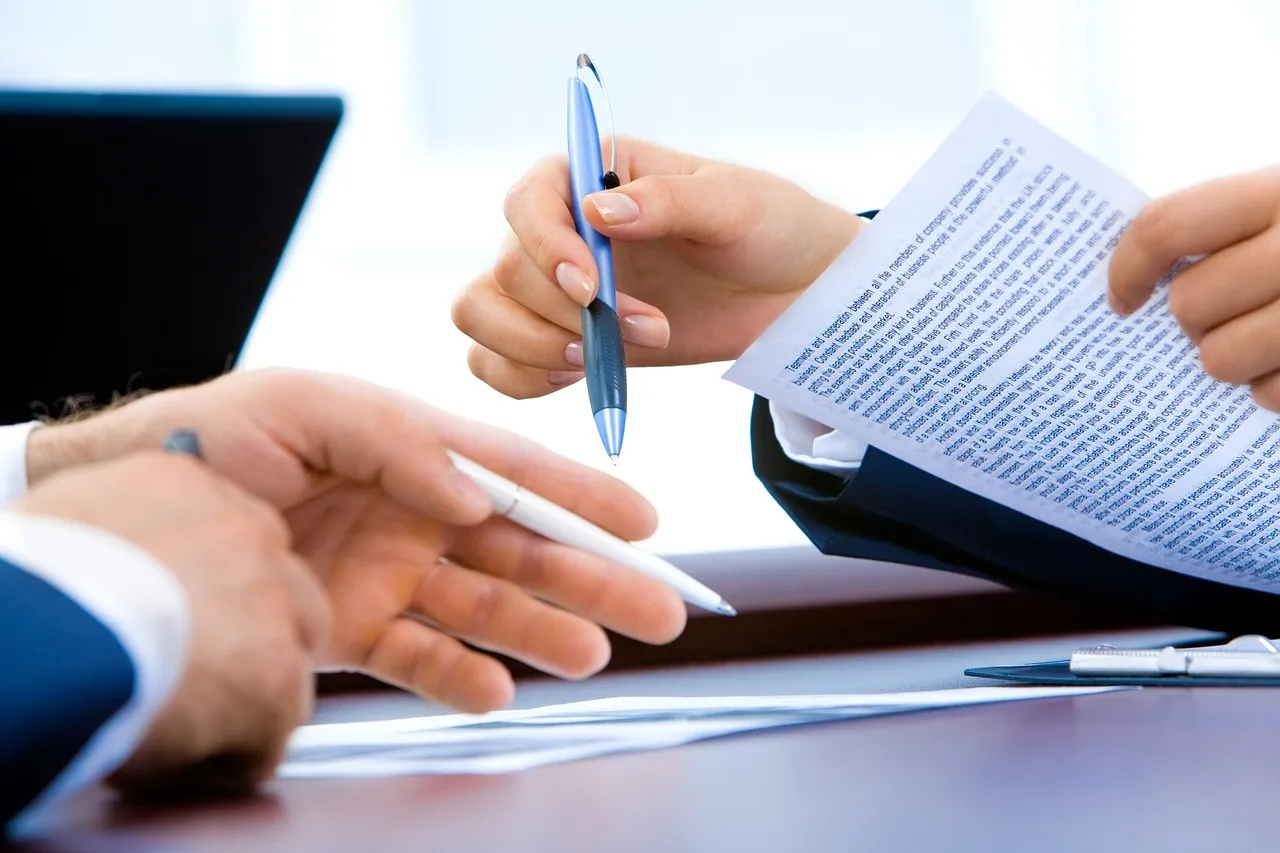 Two people reviewing and signing a contract with pens at a desk.