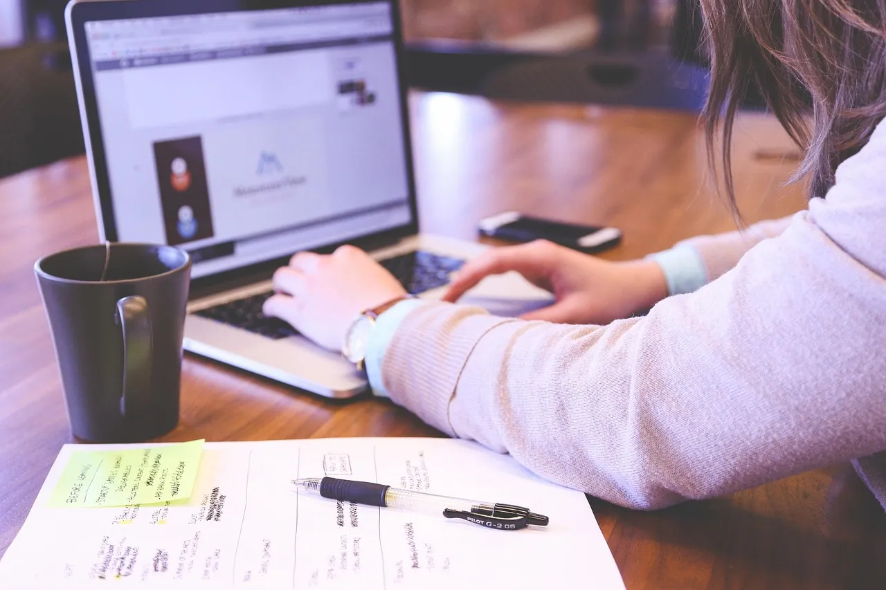 Woman in glasses working late at night on laptop, holding coffee, making phone call.
